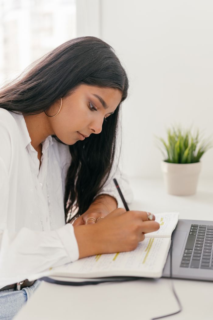 Young woman intensely writing notes in a notebook at home, engaging in online learning with a laptop.